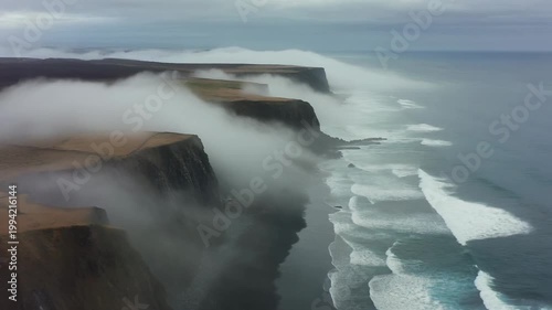 Aerial view of rugged cliffs and icy waters with fog rolling in over the ocean's edge.