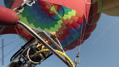 A vibrant hot air balloon with a colorful pattern is being inflated under a clear blue sky, viewed from the basket.