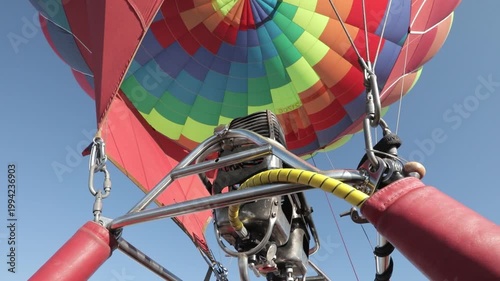 A vibrant hot air balloon with a colorful pattern is being inflated under a clear blue sky, viewed from the basket.