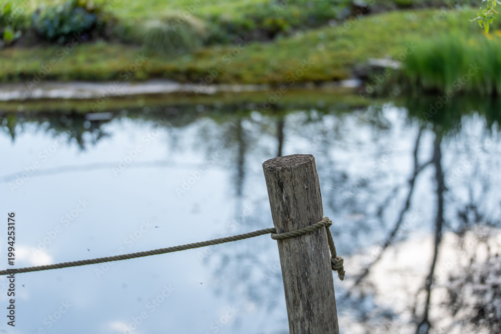 Fototapeta premium Rustic wooden post stands by calm pond water. Faint reflections ripple gently across the surface. Green grass and reeds frame the tranquil background. Weathered rope ties the post, adding rustic charm