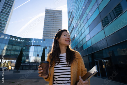 Smiling businesswoman holding coffee and laptop in modern city