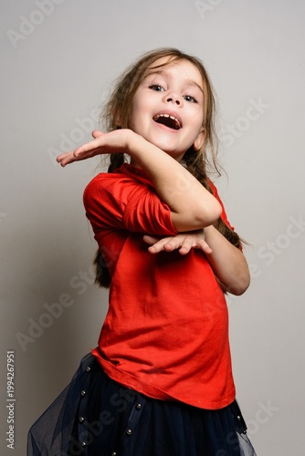 A cheerful young girl in a red shirt and dark skirt, playfully posing against a neutral background.