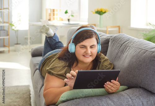 Chubby overweight young woman enjoys lazy day, lying on comfortable sofa and listening to music or watching videos via headphones and tablet, poses at home, uses modern technologies