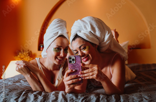 Two happy women wearing under-eye patches and towels using smartphone in bed