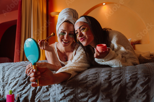 Two happy women taking selfie with under eye patches and towels on head