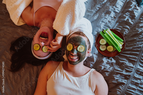 Two happy women lying on bed applying face mask and cucumber slices
