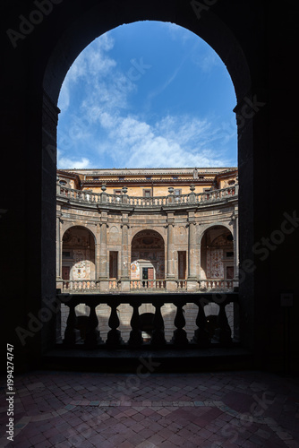 A framed view of the circular courtyard and upper loggia at Villa Farnese in Caprarola, Italy, showcasing Renaissance architecture under a blue sky.