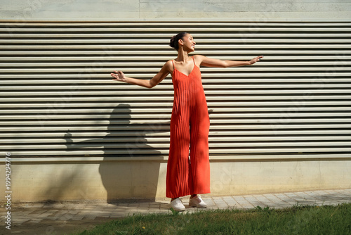 Energetic female in vivid orange clothing standing on verdant grass beside weathered metal wall