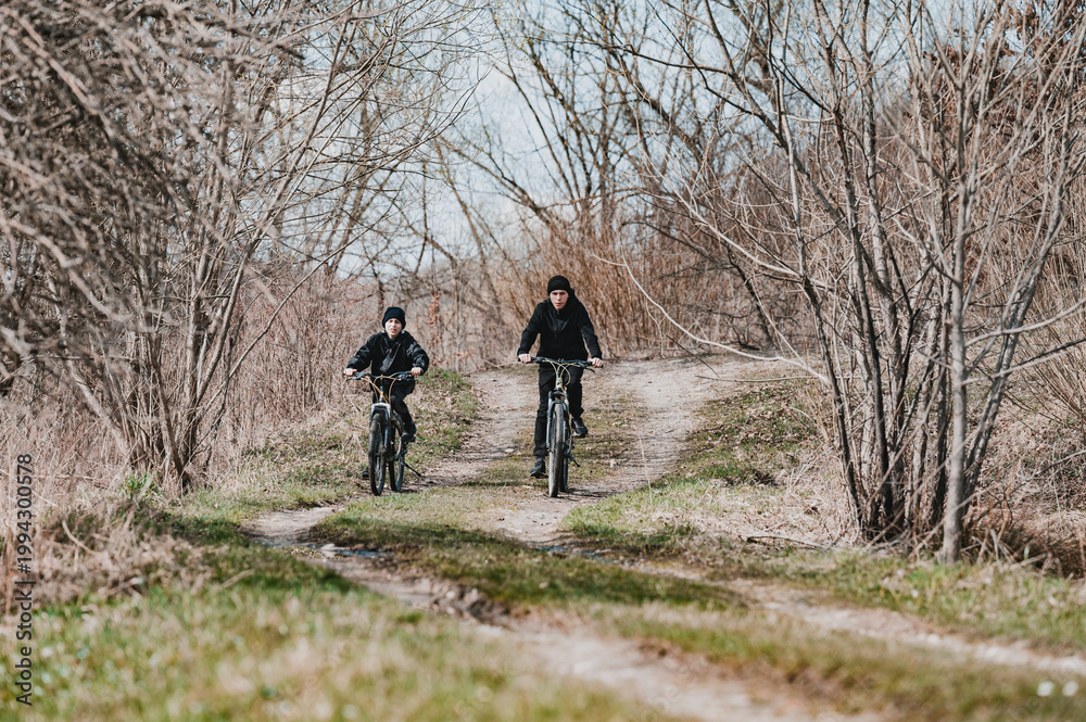 Fototapeta premium Two cyclists ride along a dirt path through a leafless winter forest. A serene and peaceful outdoor scene.
