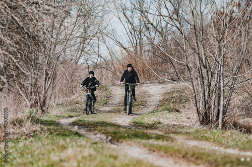 Two cyclists ride along a dirt path through a leafless winter forest. A serene and peaceful outdoor scene.