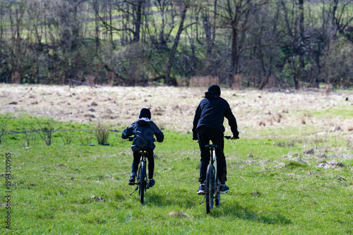 Two cyclists ride through a verdant field, trees forming a backdrop. A peaceful outdoor scene.