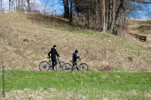 Two cyclists enjoying a sunny day, biking along a grassy path. A scenic woodland backdrop adds to the peaceful atmosphere.