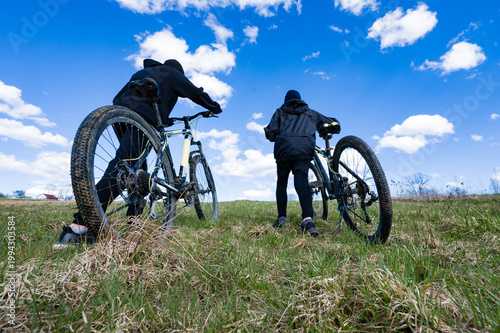 Two cyclists walk their bikes up a grassy hill under a bright blue sky. A day of adventure awaits