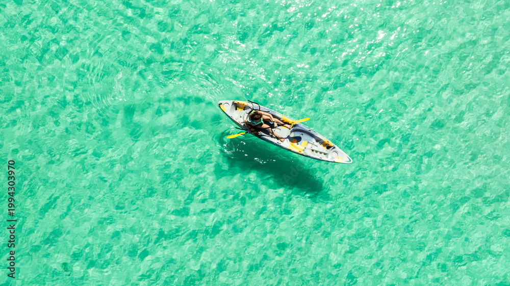 Naklejka premium High angle aerial view of a woman paddling a kayak in crystal clear turquoise ocean water near a tropical white sand beach with palm tree shadows, concept of summer luxury vacation and travel. 