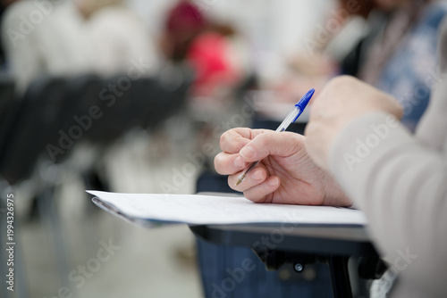 Person taking notes at a desk in a classroom or lecture hall. Close-up view of hands writing a test or essay with blurred background. Education and professional training. Photo