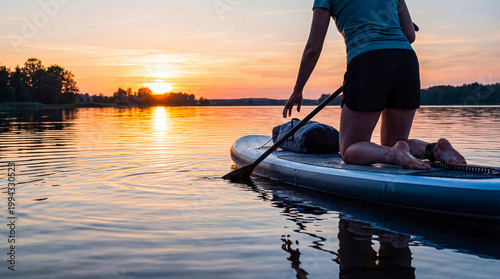 Fotografie Person kneeling on a paddleboard on a tranquil lake at sunset, enjoying a peaceful evening on the water