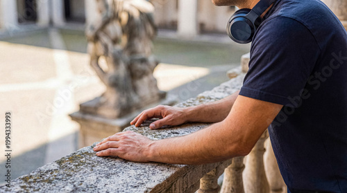 Thoughtful man with headphones around his neck, leaning on a weathered stone railing in a sunlit outdoor setting.