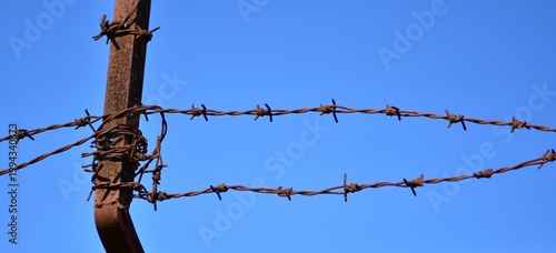 Rusty barbed wire on a vintage metal fence post against a clear blue sky. Symbolic image of border security, prison, human rights, and historical memory at Kaunas Ninth Fort memorial site.
