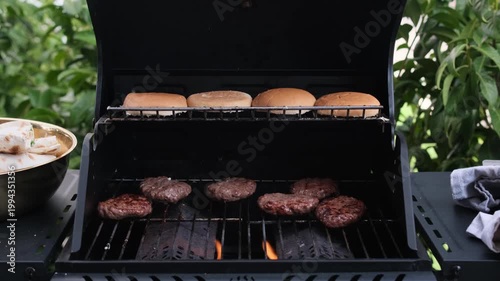 Beef burger patties grilling on barbecue with buns warming above, outdoor BBQ cooking in backyard grill