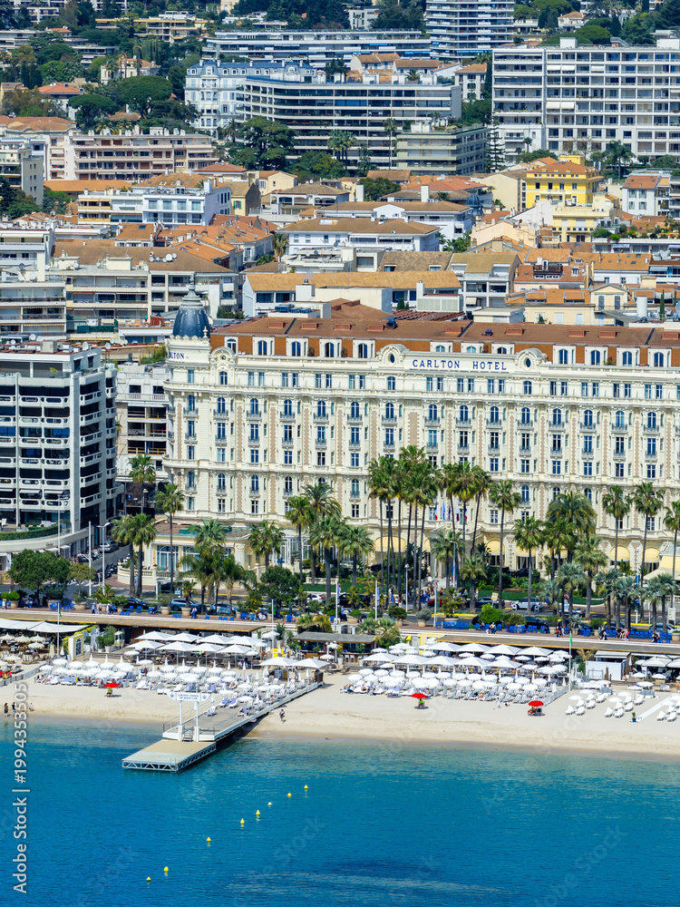 Fototapeta premium Aerial view of Cannes, France, shows Carlton Hotel facing Bay of Cannes. La Croisette lines with palms, beach clubs, sun loungers, and a pier, in crisp daylight.