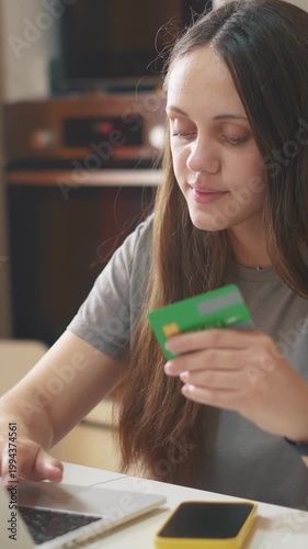 A young woman is smiling while holding a green credit card. She is using a laptop on a table, indicating an online shopping or payment activity.