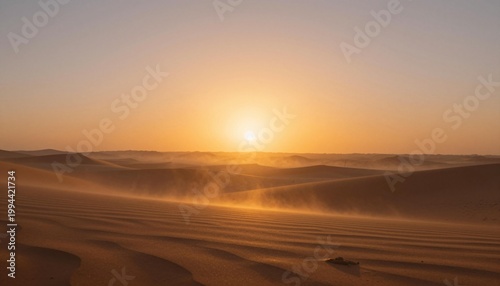 Golden desert sunrise over rippled sand dunes with drifting sand and a warm glowing sky
