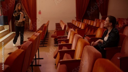 Young woman rehearsing alone in an empty theater auditorium, passionately gesturing and practicing lines between rows of red seats before a dramatic performance