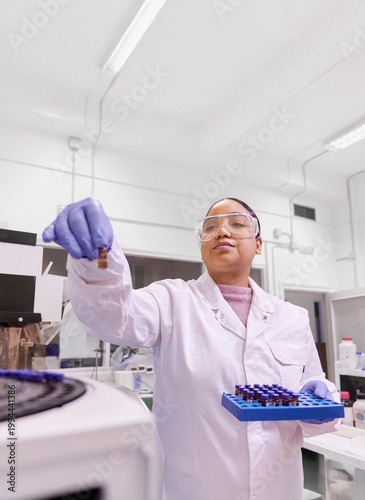 Latina researcher in lab coat and protective gear inserting sample tubes into an automatic analysis machine in a laboratory