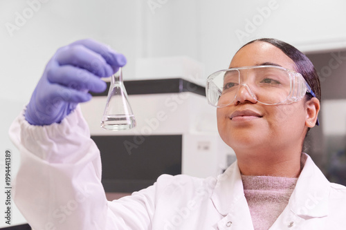 Latina female researcher smiling as she observes a biochemical sample tube in the biotech lab wearing white coat and safety glasses.