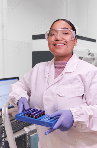 Young Dominican woman in a lab coat, gloves, and safety goggles posing in her lab holding a plastic tray of biochemical samples