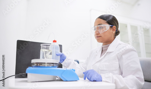 Latina scientist in a white coat operates a heating magnetic stirrer while conducting a precise chemical experiment in a research lab focused on pharmaceutical or academic innovation