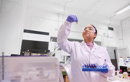Latina woman in lab coat, safety goggles, and gloves analyzing biochemical samples in a laboratory full of scientific equipment