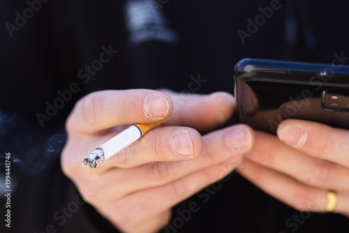 Fotografija Close-up of hands holding a smartphone and a cigarette, with smoke rising, again