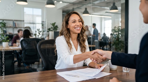 Confident businesswoman smiling and shaking hands with partner at desk in creative office, successful contract signing and human resources recruitment process concept