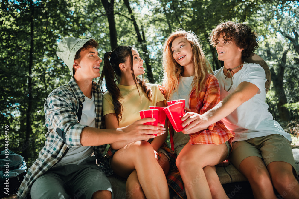 Obraz premium Four teenage friends camping in the woods sharing red cups, laughing and sitting together on a blanket during a sunny summer outdoor gathering