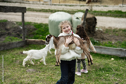 Young girl holding a baby goat on a farm smiling while playing outdoors with other children in a rural pasture