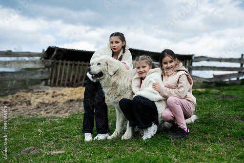 Three girls hugging a large white farm dog outdoors by a wooden fence on a cloudy rural day