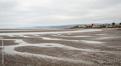 Cloudy day on a wide tidal beach with rippled sand patterns, shallow pools of water and distant houses along the muted coastline under a grey sky

