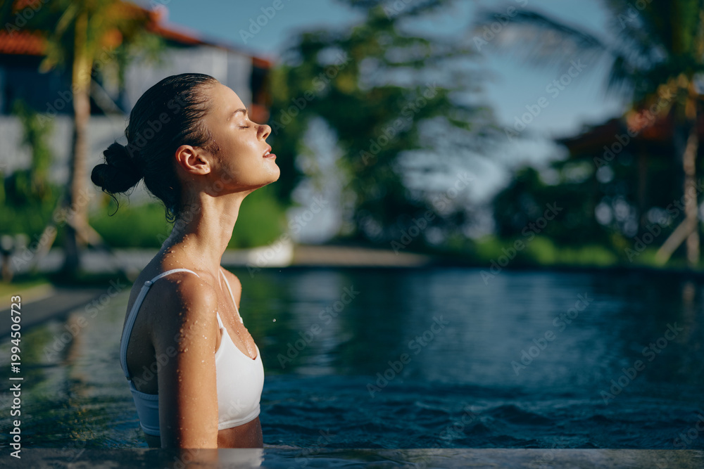 Fototapeta premium A relaxed woman stands in a sunlit pool, eyes closed, savoring the warm breeze. A casual lifestyle moment that conveys calm, self care, and a peaceful, rejuvenating escape.