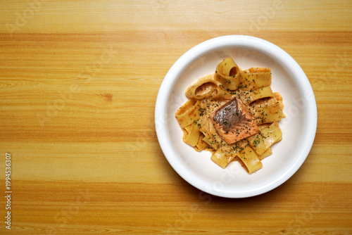 Top view of salmon pasta on white plate over wooden table, creamy sauce coating wide noodles with grilled fish. Minimal food styling, warm tones, gourmet Italian cuisine concept, restaurant menu.
