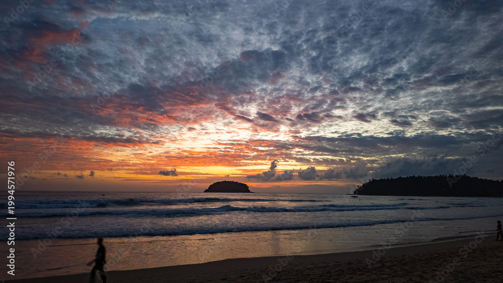 Αυτοκόλλητο A breathtaking view of the sun setting over the ocean, casting warm golden hues across the waves and clouds while people enjoy the tranquil shoreline