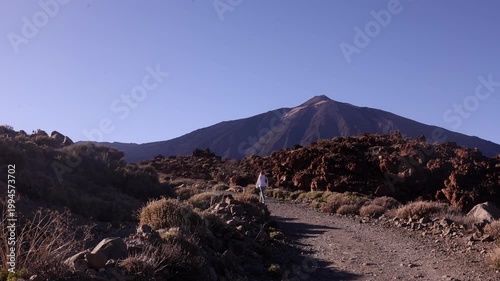 Wide cinematic shot of lone woman in white standing small against massive Mount Teide volcano and dramatic red lava field in Tenerife under deep blue sky. Epic scale travel footage.