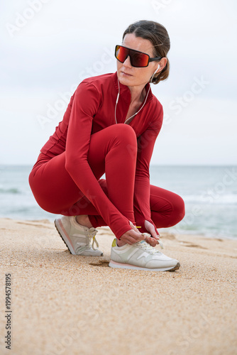 Woman In Red Activewear Tying Running Shoes On Beach Before Outdoor Workout