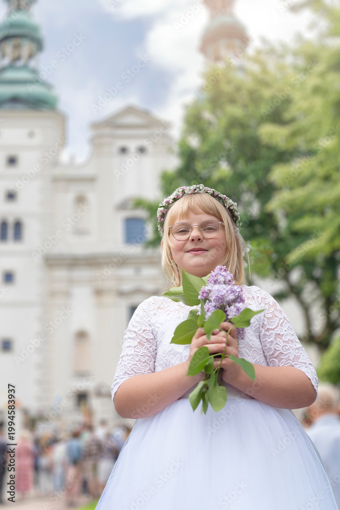 Fototapeta premium First communion celebration in Poland. Happy blonder girl in white dress, flower wreath and glasses