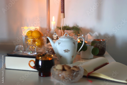 Cup of tea or coffee, teapot, book, notebook, e-reader, glasses, cookies, nuts, lemons, house plants and lit candles on the table. Hygge at home. Selective focus.