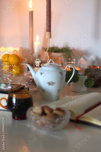 Cup of tea or coffee, teapot, book, notebook, e-reader, glasses, cookies, nuts, lemons, house plants and lit candles on the table. Hygge at home. Selective focus.