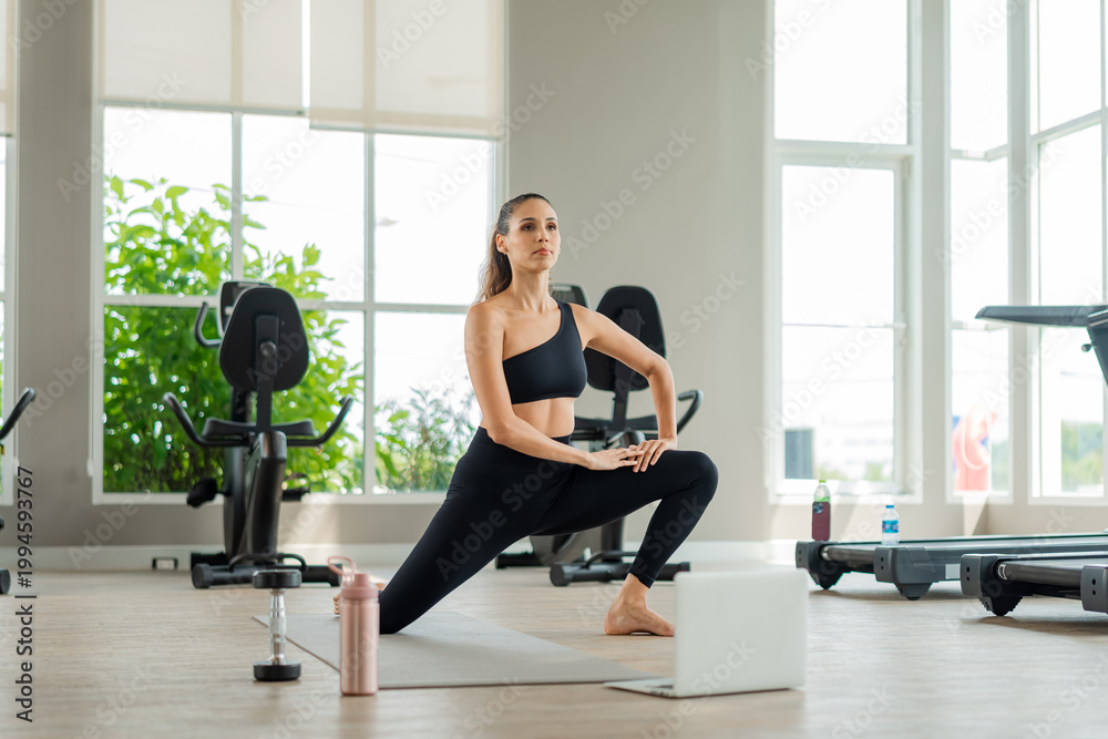 Fototapeta premium Young woman doing yoga stretch inside well-lit fitness studio. Female athlete doing yoga and fitness exercises with virtual class on computer, digital health and active lifestyle concept.