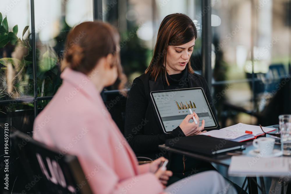 Fototapeta premium Woman presenting tablet with data charts to a colleague during a casual business meeting. Colleagues discuss financial graphs and notes at a cafe table while reviewing reports.