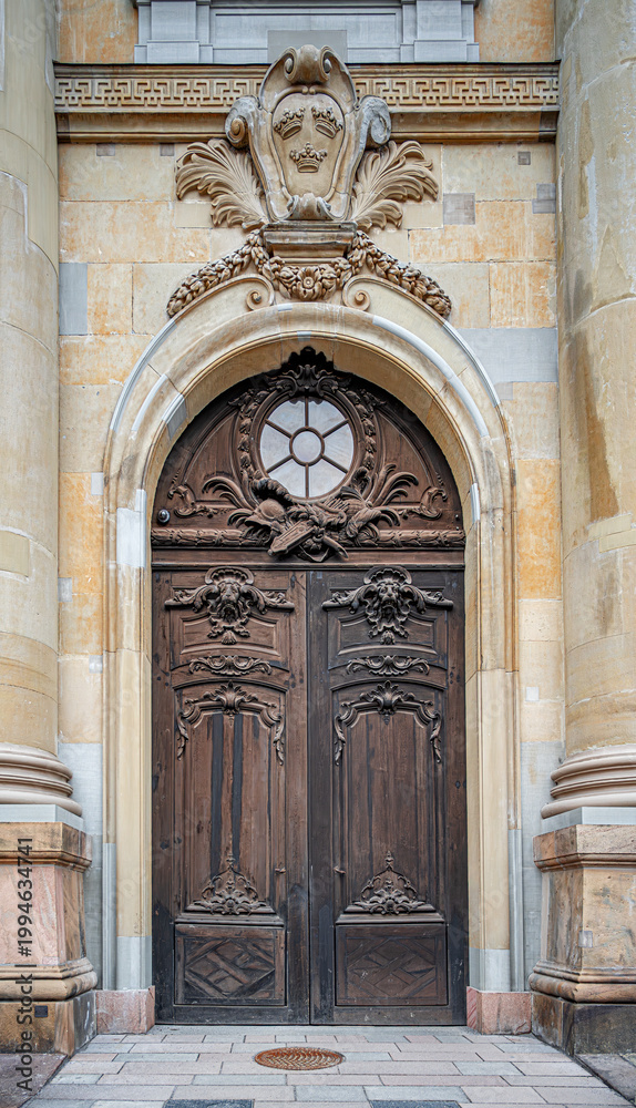 Obraz premium A grand ornate wooden entrance door with intricate carvings and a stone archway is set into the historical facade of a building in Stockholm, Sweden, featuring the iconic three crowns heraldic symbol.
