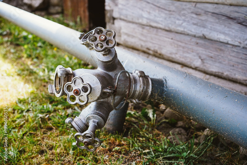 Industrial metal pipe assembly with multiple control valves and handles outdoors on grass next to a wooden building wall.